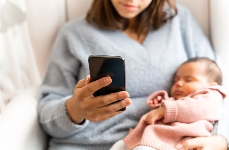 Mother holding her newborn baby in her arms while checking her phone, calm and quiet moment at home